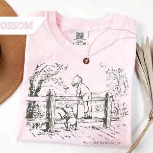 May include: A light pink t-shirt with a black and white illustration of a scene from Hundred Acre Wood. The shirt features a crew neck and the text "Hundred Acre Wood" at the bottom. A gold necklace with a pendant is on the shirt.