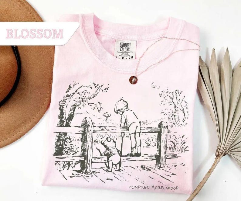 May include: A light pink t-shirt with a black and white illustration of a scene from Hundred Acre Wood. The shirt features a crew neck and the text "Hundred Acre Wood" at the bottom. A gold necklace with a pendant is on the shirt.
