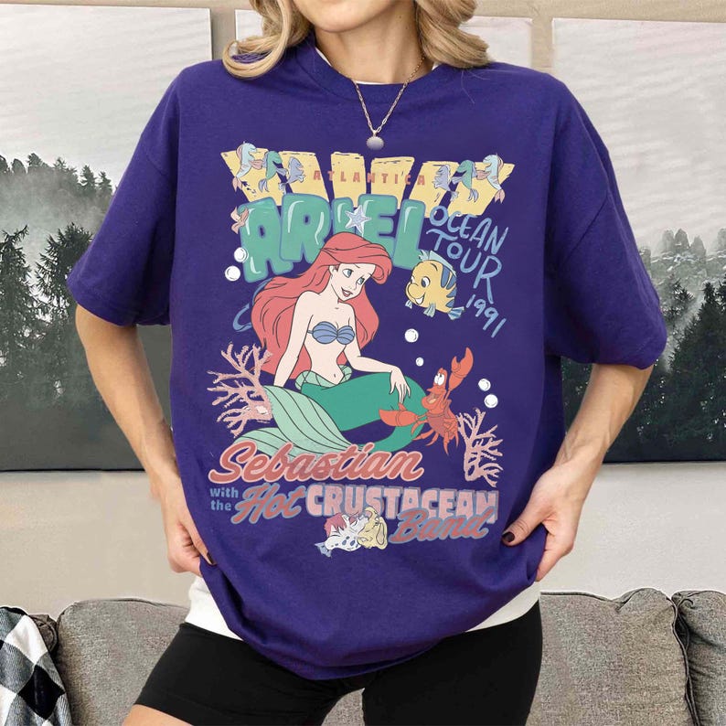 K&ouml;nnte beinhalten: Ein lila T-Shirt mit einem Motiv von Arielle, der Meerjungfrau. Die Grafik zeigt Arielle, Fabius, Sebastian und den Text "Ariel Ocean Tour 1991" und "Sebastian with the Hot Crustacean Band."