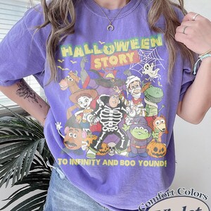May include: A violet Comfort Colors t-shirt with a Halloween-themed graphic. The text reads "HALLOWEEN STORY" and "TO INFINITY AND BOO YOUND!" The graphic includes cartoon characters, pumpkins, and a spiderweb.