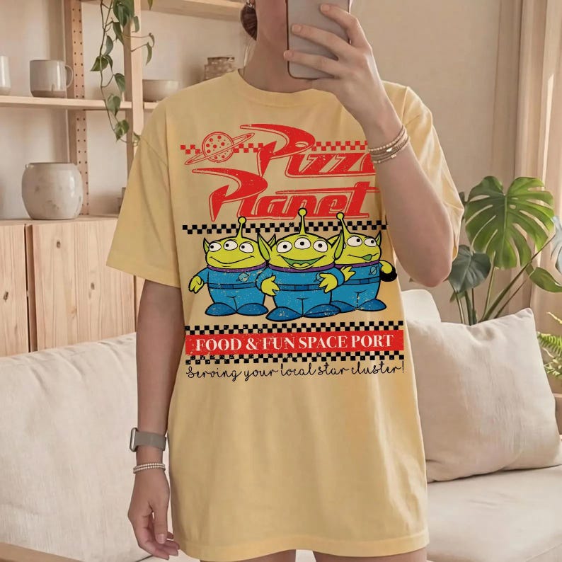 May include: A pale yellow t-shirt featuring a red Pizza Planet logo with a planet graphic, three green three-eyed aliens in blue uniforms, and the text FOOD & FUN SPACE PORT Serving your local star cluster in a black and white checkered frame.