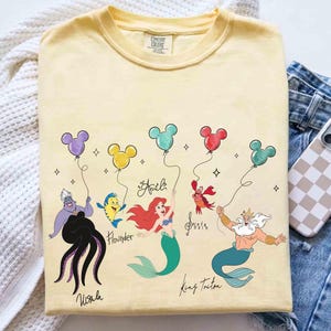 Puede incluir: Camiseta amarilla Comfort Colors con personajes de La Sirenita de Disney sosteniendo globos de Mickey Mouse. Los personajes incluyen a &Uacute;rsula, Flounder, Ariel, Sebasti&aacute;n y el Rey Trit&oacute;n. Los nombres est&aacute;n escritos debajo.
