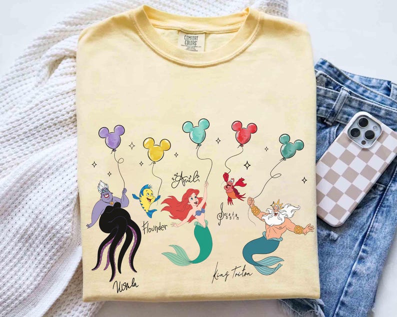 Puede incluir: Camiseta amarilla Comfort Colors con personajes de La Sirenita de Disney sosteniendo globos de Mickey Mouse. Los personajes incluyen a &Uacute;rsula, Flounder, Ariel, Sebasti&aacute;n y el Rey Trit&oacute;n. Los nombres est&aacute;n escritos debajo.