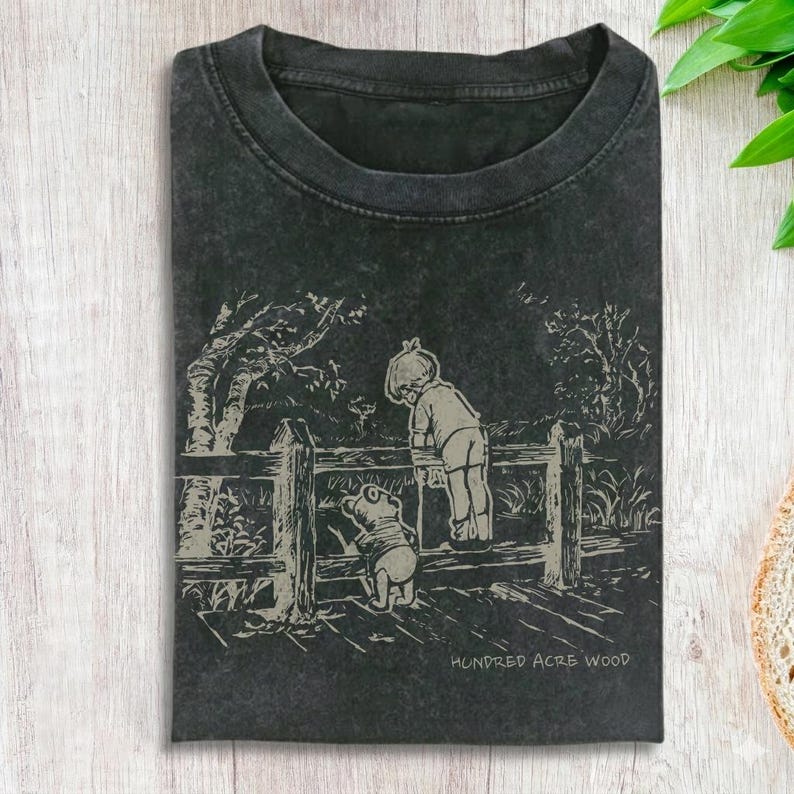 May include: Dark gray t-shirt featuring a black and white illustration of a boy and a bear looking over a wooden fence. The text "Hundred Acre Wood" is printed at the bottom of the graphic. The shirt has a crew neck and short sleeves.