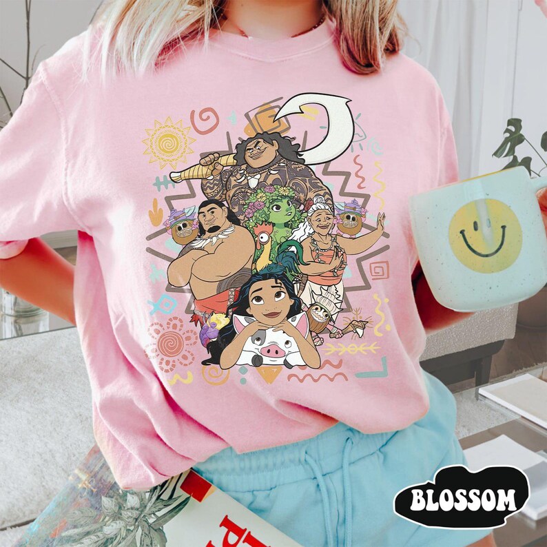 May include: A light pink t-shirt featuring a colorful graphic print of characters from the movie Moana, including Moana, Maui, Pua, Heihei, and Gramma Tala, surrounded by various tribal-inspired shapes, symbols, and patterns in yellow, orange, and teal.