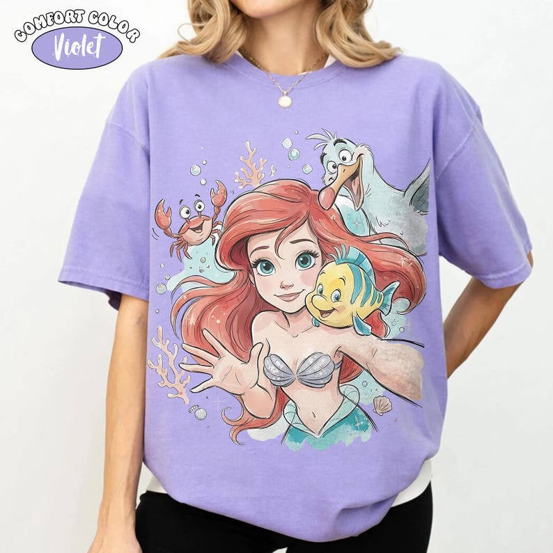 May include: A violet t-shirt featuring a graphic print of a red-haired mermaid, a yellow and blue fish, a small red crab, and a white seagull. The text COMFORT COLOR Violet is printed in the upper left corner above the graphic illustration.