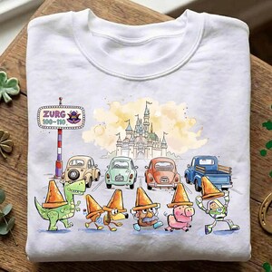 May include: White sweatshirt featuring a colorful cartoon design. The design includes a castle, cars, and characters wearing orange traffic cones. The text "ZURG 100-110" is also visible. The overall theme is playful and whimsical.