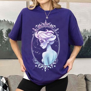 May include: A purple t-shirt featuring a stylized illustration of a woman with white hair and a blue complexion, framed within an oval border. The design includes snowflake and star accents. The shirt has short sleeves.
