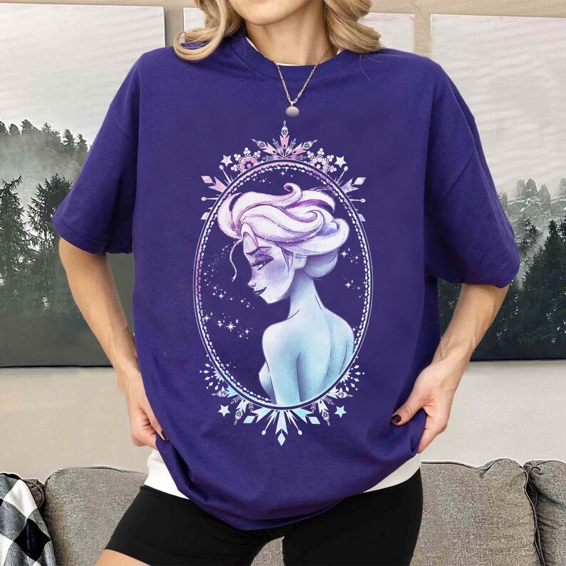 May include: A purple t-shirt featuring a stylized illustration of a woman with white hair and a blue complexion, framed within an oval border. The design includes snowflake and star accents. The shirt has short sleeves.