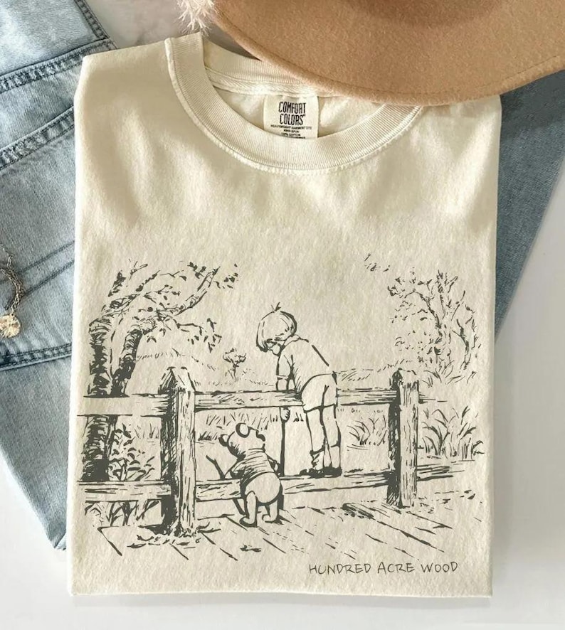 May include: Cream-colored t-shirt featuring a black and white illustration of a boy and a bear looking over a wooden fence. The text "Hundred Acre Wood" is printed at the bottom. The shirt has a Comfort Colors tag.