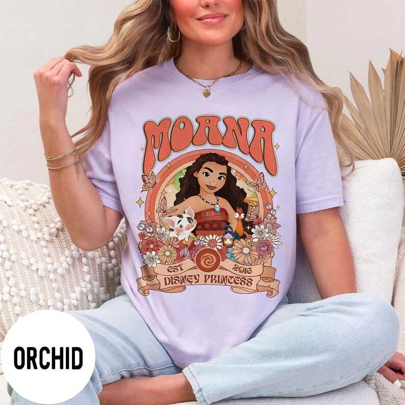 Op de afbeelding: Lichtpaars T-shirt met het woord "MOANA" in oranje retro letters. Het shirt heeft een afbeelding van Moana, Pua, Hei Hei en bloemen. De tekst "EST 2016 DISNEY PRINCESS" staat ook op het shirt.