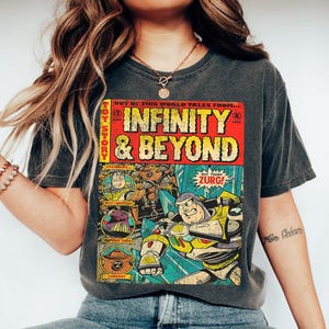Może przedstawiać: Ciemnoszara koszulka z grafiką w stylu komiksu. Grafika przedstawia napis "INFINITY & BEYOND" oraz wizerunki Buzza Astrala i Zurga. Koszulka ma wygląd vintage.