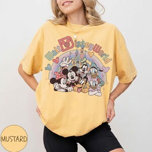 May include: Mustard yellow t-shirt featuring a colorful graphic of Disney characters, including Mickey and Minnie Mouse, Goofy, Donald Duck, and Pluto. The text "Walt Disney World" arches above the characters, with a rainbow and castle in the background.