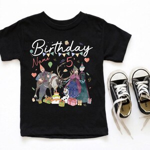 May include: Black short-sleeved t-shirt with a colorful graphic featuring characters and the text "Birthday Name 5". The graphic includes a cake, presents, and a banner. Black canvas sneakers with white laces are next to the shirt.