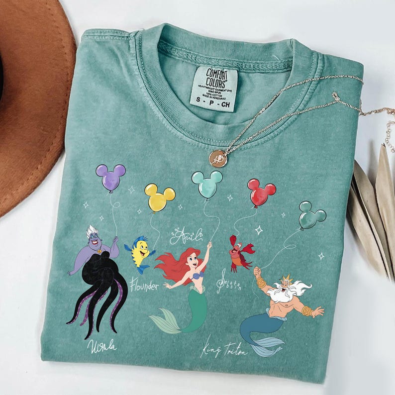 Puede incluir: Una camiseta Comfort Colors turquesa con un gr&aacute;fico de Ariel, Flounder, Sebasti&aacute;n, el Rey Trit&oacute;n y &Uacute;rsula, cada uno sosteniendo un globo de Mickey Mouse. La camiseta tambi&eacute;n tiene el nombre "Ariel" escrito en cursiva. Un collar plateado con un colgante est&aacute; sobre la camiseta.