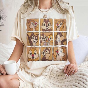 May include: Cream-colored t-shirt featuring a grid of nine cartoon chipmunks in various poses. The chipmunks are in brown squares with a light brown background. The shirt has short sleeves and a relaxed fit.