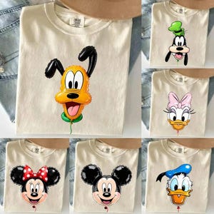 May include: A collection of cream-coloured crew neck t-shirts, each featuring a different Disney character depicted as a shiny, inflated balloon. The characters include Pluto, Goofy, Daisy Duck, Minnie Mouse, Mickey Mouse, and Donald Duck.