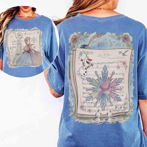 Pode incluir: Camiseta azul clara com um gr&aacute;fico de Frozen na frente. A parte de tr&aacute;s mostra partituras, Olaf e um desenho de floco de neve. O texto "Let It Go" tamb&eacute;m &eacute; vis&iacute;vel.