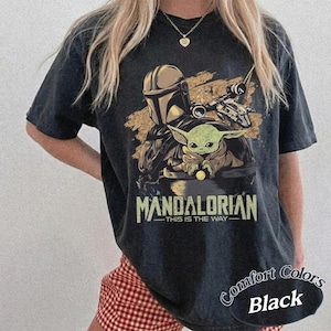 May include: Black t-shirt featuring a graphic of The Mandalorian and Grogu, with a spaceship in the background. The text "MANDALORIAN" and "THIS IS THE WAY" are printed below the image. The shirt has a distressed look and the words "Comfort Colors Black" are printed on the bottom right.
