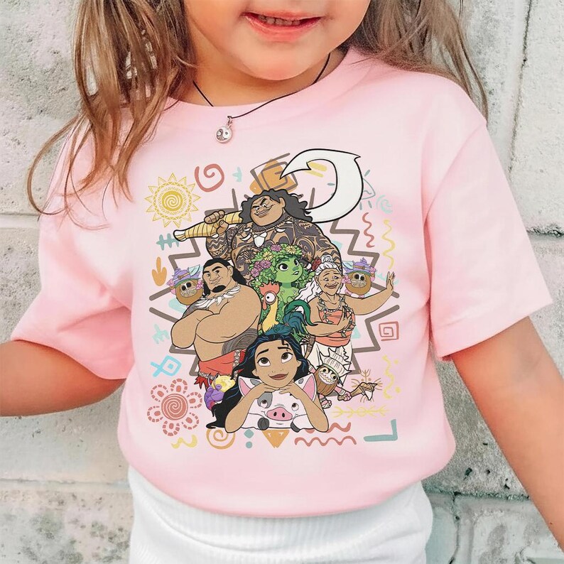 May include: A light pink short-sleeved t-shirt featuring a colorful graphic print of characters from the movie Moana, including Moana, Maui, Pua, Heihei, and Gramma Tala, surrounded by various tribal-inspired symbols and patterns.