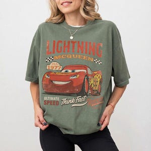 Puede incluir: Camiseta verde con Lightning McQueen de la pel&iacute;cula Cars. El dise&ntilde;o incluye el texto "LIGHTNING MCQUEEN" en naranja, el a&ntilde;o "1977" y las frases "ULTIMATE SPEED" y "Think Fast". El gr&aacute;fico es de estilo vintage.