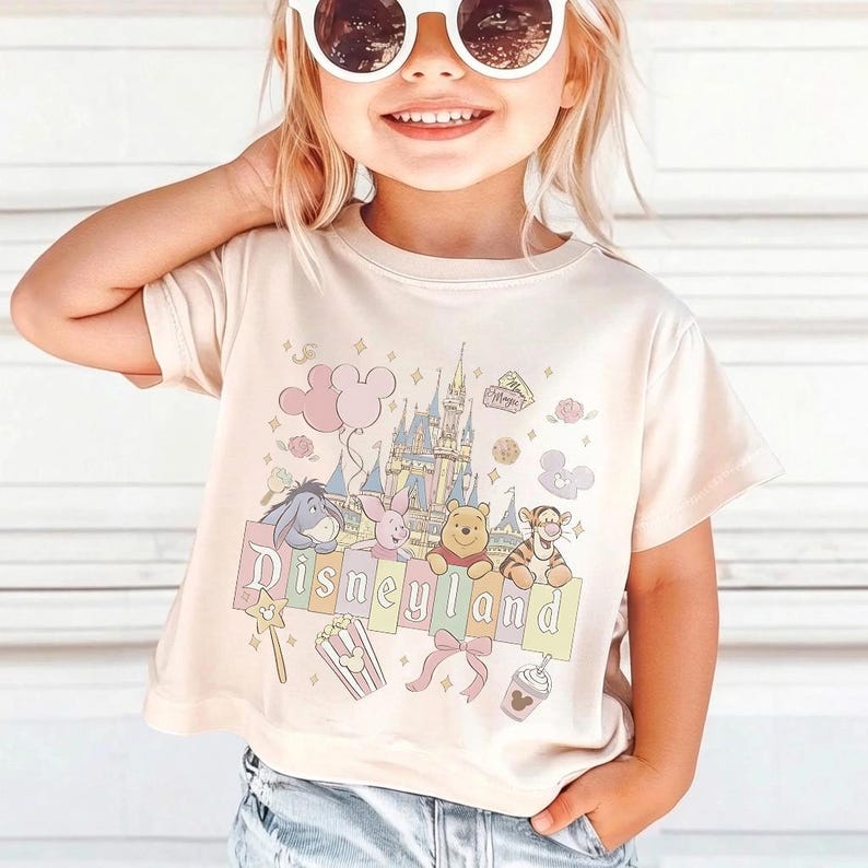 May include: A light beige t-shirt with the word "Disneyland" in pastel colours, featuring cartoon characters and a castle graphic. The shirt has short sleeves and is worn by a young child wearing sunglasses.