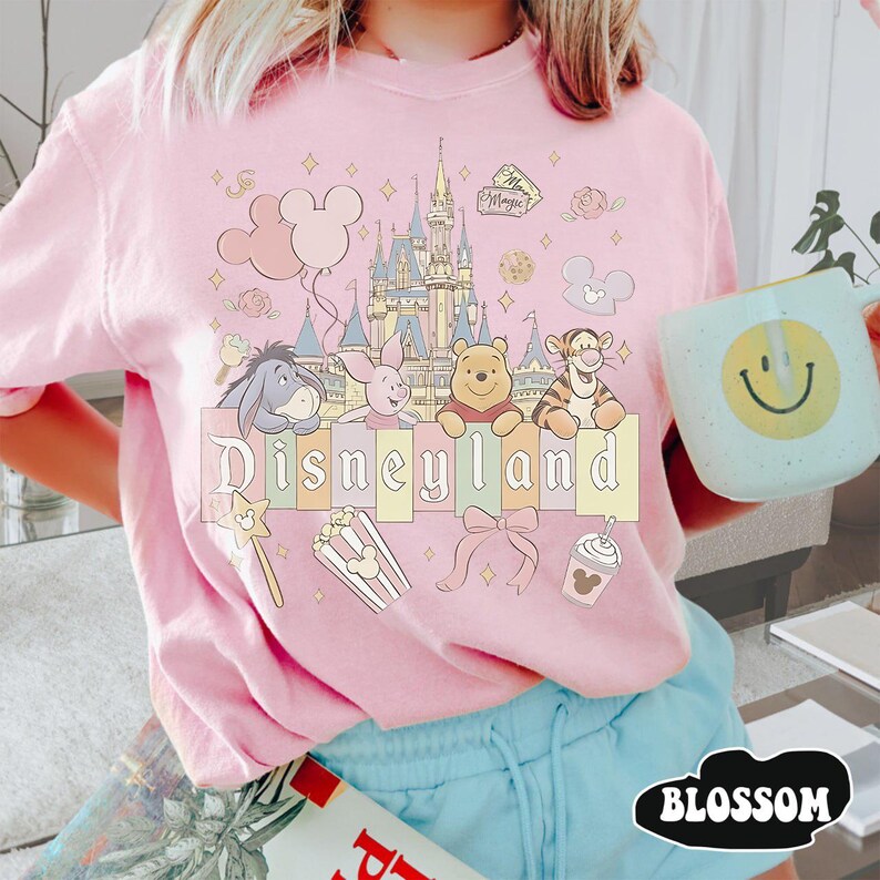 May include: Pale pink Disneyland t-shirt with a colourful graphic of Disney characters, a castle, and the word "Disneyland". The shirt includes images of balloons, popcorn, and a bow. A light blue mug with a smiley face is held in the image.