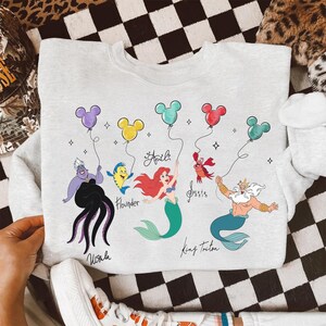 Puede incluir: Una sudadera gris claro con un gr&aacute;fico colorido de personajes de La Sirenita. El dise&ntilde;o incluye a &Uacute;rsula, Flounder, Ariel, Sebasti&aacute;n y el Rey Trit&oacute;n, cada uno sosteniendo un globo con forma de Mickey Mouse. Los nombres de los personajes est&aacute;n escritos debajo.