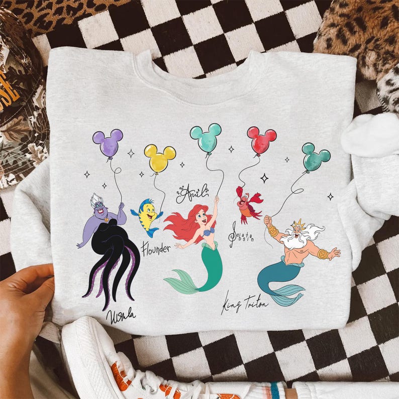 Puede incluir: Una sudadera gris claro con un gr&aacute;fico colorido de personajes de La Sirenita. El dise&ntilde;o incluye a &Uacute;rsula, Flounder, Ariel, Sebasti&aacute;n y el Rey Trit&oacute;n, cada uno sosteniendo un globo con forma de Mickey Mouse. Los nombres de los personajes est&aacute;n escritos debajo.