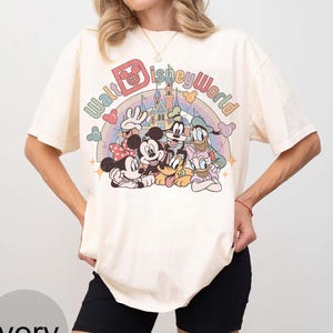 May include: Ivory-colored t-shirt featuring a colorful graphic of Disney characters, including Mickey and Minnie Mouse, Donald Duck, and Goofy. The text "Walt Disney World" is arched above the characters. The shirt has short sleeves and a relaxed fit.