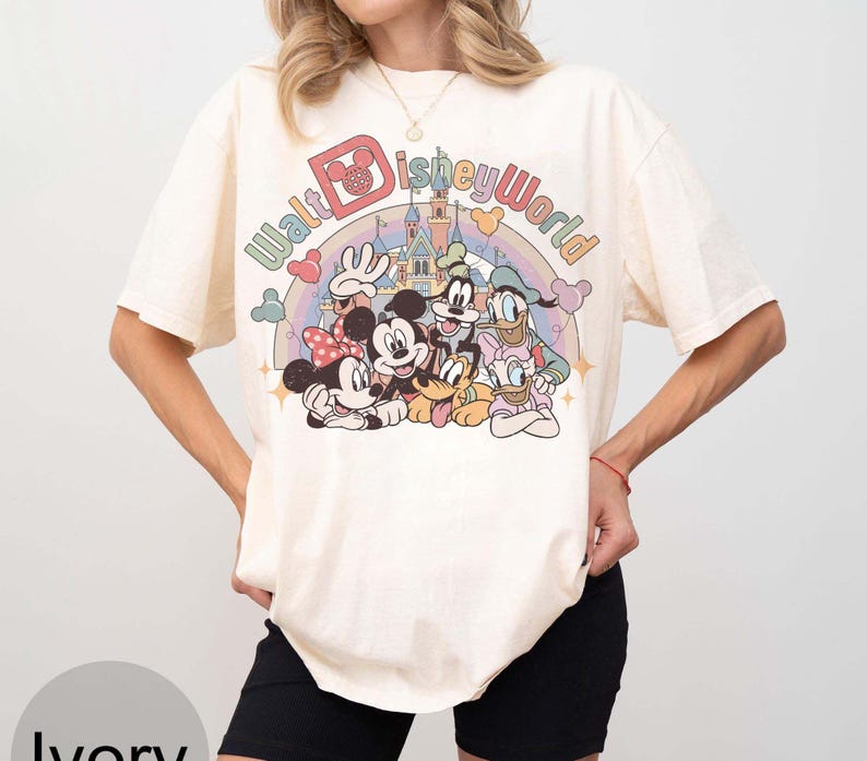 May include: Ivory-colored t-shirt featuring a colorful graphic of Disney characters, including Mickey and Minnie Mouse, Donald Duck, and Goofy. The text "Walt Disney World" is arched above the characters. The shirt has short sleeves and a relaxed fit.