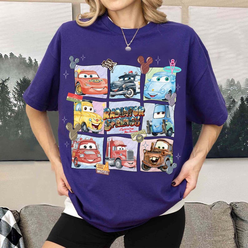 May include: A purple t-shirt featuring a grid of cartoon cars from the movie "Cars." The design includes characters like Lightning McQueen and Mater, with the words "Radiator Springs" prominently displayed. The shirt has a relaxed fit.