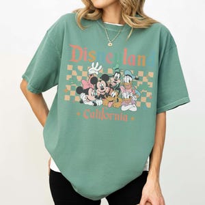 May include: Green t-shirt featuring the text "Disneyland California" with a graphic of Mickey Mouse, Minnie Mouse, Goofy, Donald Duck, and Daisy Duck. The shirt has a crew neck and short sleeves.