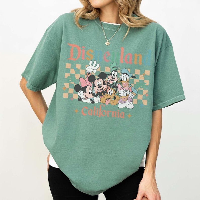 May include: Green t-shirt featuring the text "Disneyland California" with a graphic of Mickey Mouse, Minnie Mouse, Goofy, Donald Duck, and Daisy Duck. The shirt has a crew neck and short sleeves.