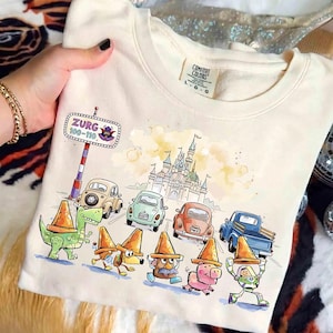 May include: Cream-colored sweatshirt featuring a colorful graphic of cartoon characters and cars, with a castle in the background. The text "Zurg 100-110" is on a sign. The sweatshirt has a Comfort Colors tag.