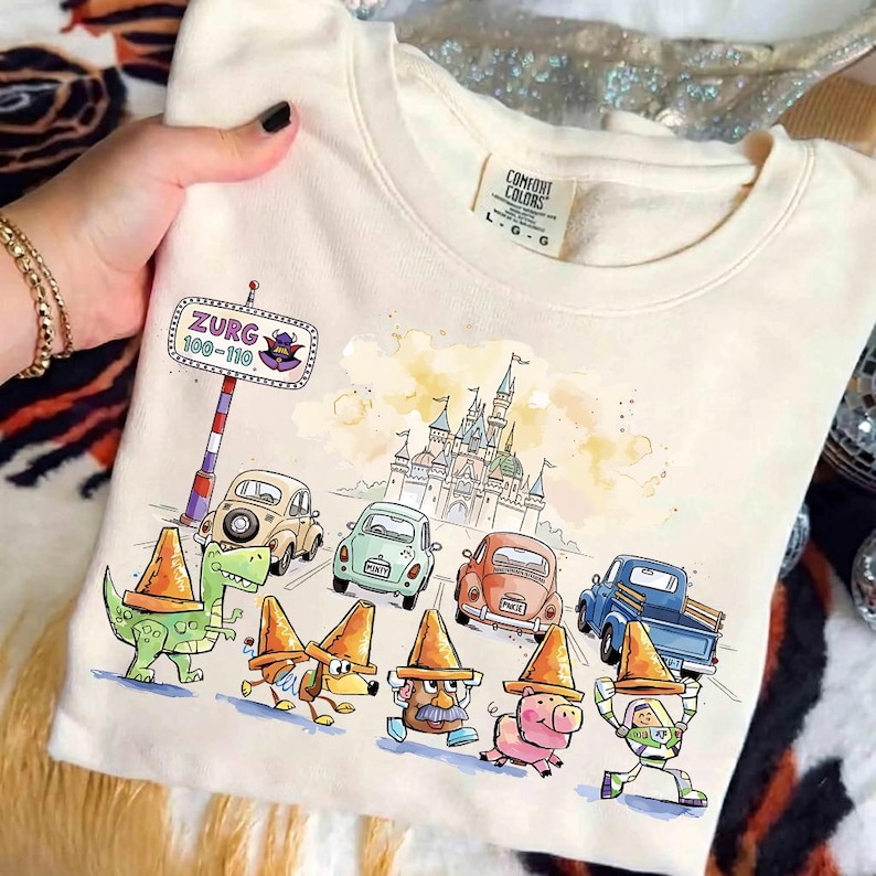 May include: Cream-colored sweatshirt featuring a colorful graphic of cartoon characters and cars, with a castle in the background. The text "Zurg 100-110" is on a sign. The sweatshirt has a Comfort Colors tag.