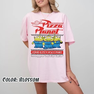 May include: A light pink t-shirt featuring a graphic print with the text Pizza Planet, Food & Fun Space Port, and Serving your local star cluster. The design includes three green three-eyed aliens in blue space suits and a checkered border.