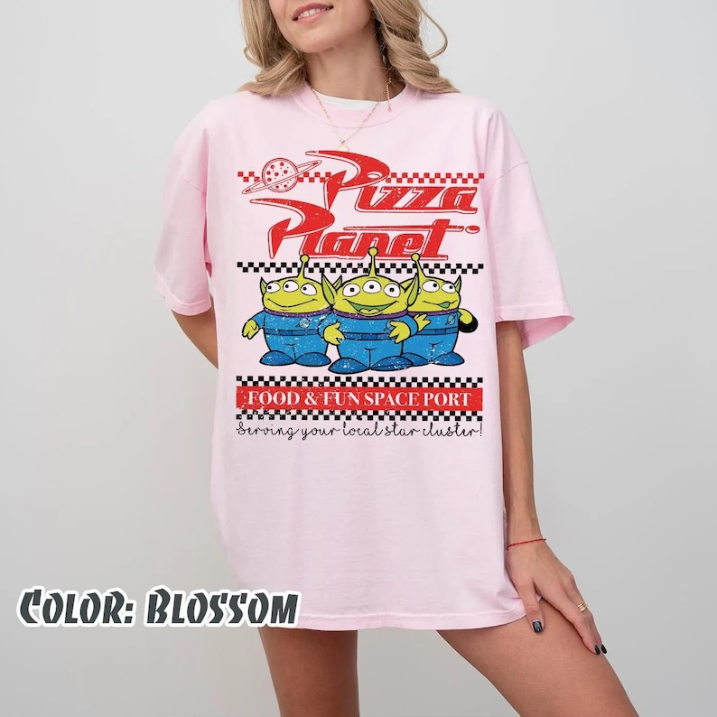 May include: A light pink t-shirt featuring a graphic print with the text Pizza Planet, Food & Fun Space Port, and Serving your local star cluster. The design includes three green three-eyed aliens in blue space suits and a checkered border.