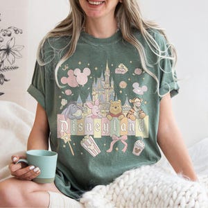 May include: Green t-shirt with a Disneyland graphic. The design features the Disneyland castle, Mickey Mouse balloons, and characters like Winnie the Pooh and Tigger. The word "Disneyland" is printed across the front in pastel colours.