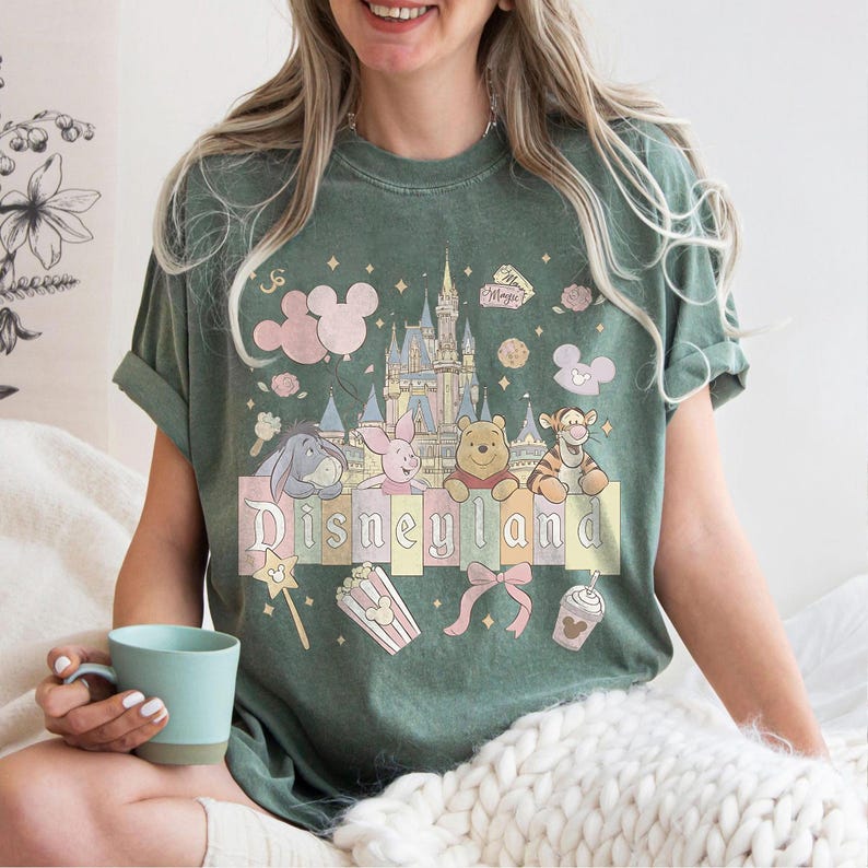 May include: Green t-shirt with a Disneyland graphic. The design features the Disneyland castle, Mickey Mouse balloons, and characters like Winnie the Pooh and Tigger. The word "Disneyland" is printed across the front in pastel colours.