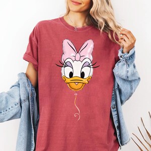 May include: A brick red short-sleeve t-shirt featuring a graphic of a Daisy Duck head shaped like a balloon. The character has a large pink bow, long black eyelashes, and a yellow beak. A thin pink string hangs from the bottom of the balloon graphic.