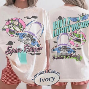 May include: An ivory t-shirt featuring Buzz Lightyear graphics. The front displays Buzz in a pink circle, a spaceship, and the text Space Ranger. The back shows the name Buzz Lightyear, a spaceship, Buzz in a circle, and To Infinity & Beyond.