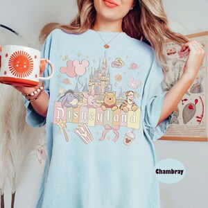 May include: Pale blue t-shirt featuring a Disneyland graphic. The design includes the Disneyland castle, Winnie the Pooh characters, balloons, and the word "Disneyland". The shirt is made of a soft, comfortable material.