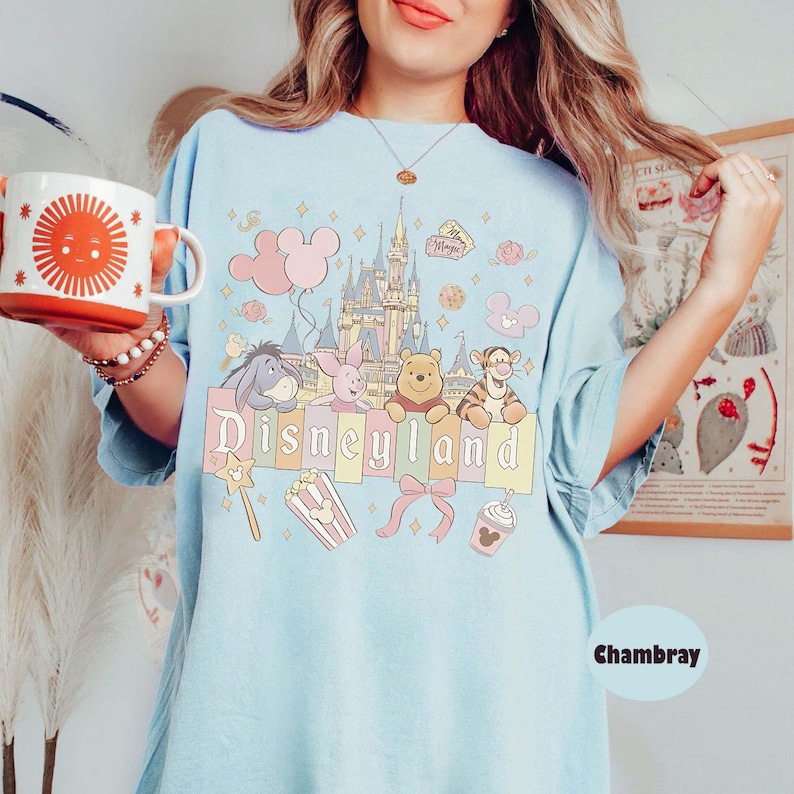 May include: Pale blue t-shirt featuring a Disneyland graphic. The design includes the Disneyland castle, Winnie the Pooh characters, balloons, and the word "Disneyland". The shirt is made of a soft, comfortable material.