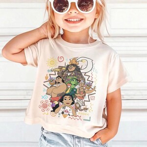 May include: A cream-colored short-sleeve t-shirt featuring a colorful graphic print of various animated characters, including a large figure with a hook, a smaller figure with a pig, and other island-themed icons like suns and waves in a central design.