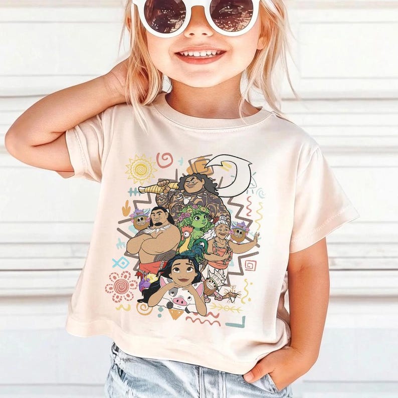 May include: A cream-colored short-sleeve t-shirt featuring a colorful graphic print of various animated characters, including a large figure with a hook, a smaller figure with a pig, and other island-themed icons like suns and waves in a central design.