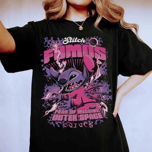Pode incluir: Camiseta preta com um desenho de Stitch de Lilo & Stitch. O design inclui as palavras "Stitch Famous" e "Fear of Missing Outer Space" em rosa e roxo, com raios.