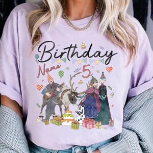 May include: A light purple t-shirt with the word "Birthday" in black script, a number "5", and cartoon characters. The design includes a birthday cake, balloons, and gift boxes. The shirt is a crew neck.