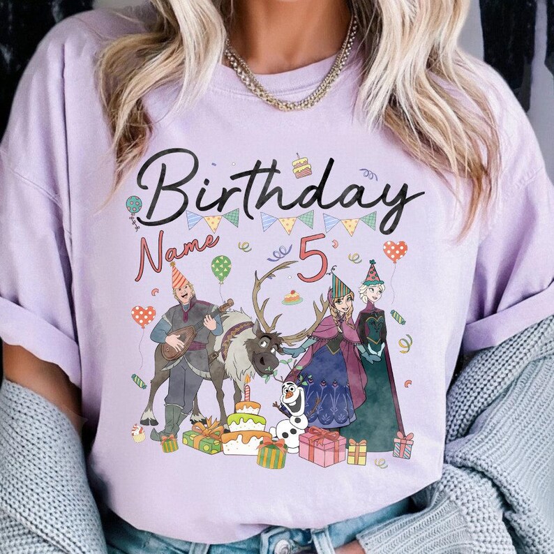 May include: A light purple t-shirt with the word "Birthday" in black script, a number "5", and cartoon characters. The design includes a birthday cake, balloons, and gift boxes. The shirt is a crew neck.