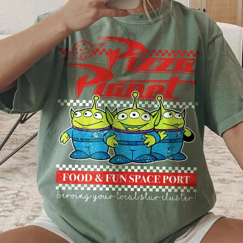 May include: A sage green t-shirt featuring a red Pizza Planet logo, three green three-eyed aliens in blue space suits, and the text FOOD & FUN SPACE PORT Serving your local star cluster, accented with red and white checkered border patterns.